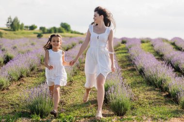 cheerful woman and girl in white dresses holding hands and running in field
