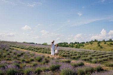 woman and girl in white dresses and straw hats walking in flowering meadow under blue sky