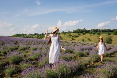 woman in straw hat and white dress walking with daughter in meadow