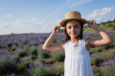 girl smiling at camera while adjusting straw hat in blurred lavender field