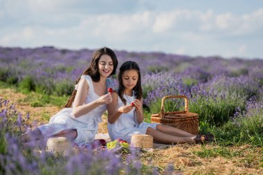 smiling mother and child eating strawberries during picnic in lavender meadow