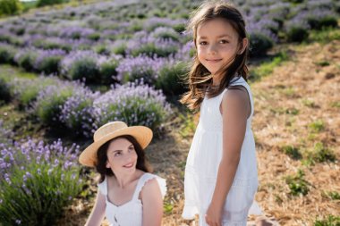 happy girl smiling at camera near blurred mom in lavender meadow