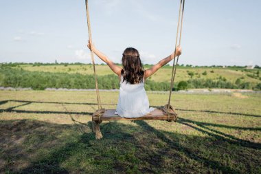 back view of child in summer dress riding swing in meadow