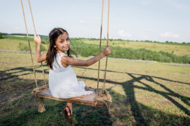 girl in white dress riding swing and looking at camera in meadow