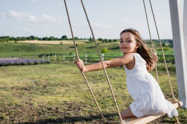 girl in summer dress looking at camera while riding swing in field