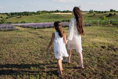 full length of mother and daughter in white dresses walking in field 