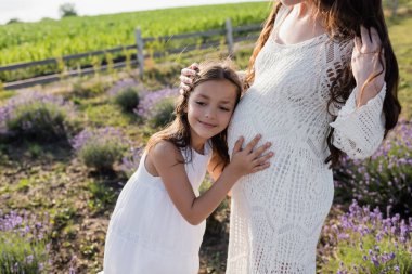 smiling child hugging belly of pregnant mother in blurred countryside field