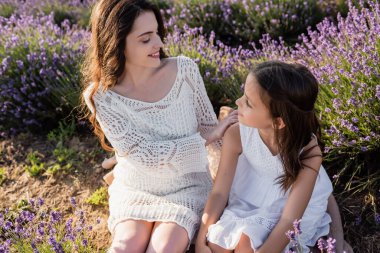 happy woman looking at daughter while sitting in meadow with blooming lavender