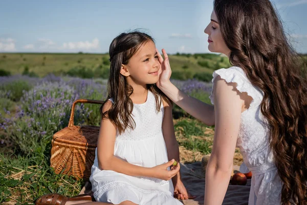 happy woman touching face of daughter while having picnic in meadow