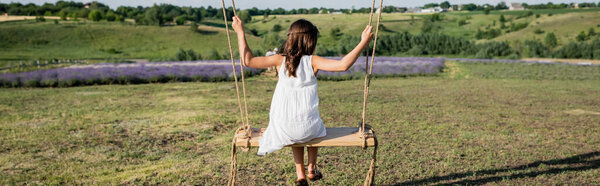 back view of kid in white summer dress riding swing in meadow, banner
