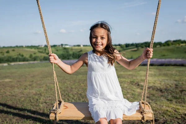 excited girl in summer dress riding swing in meadow
