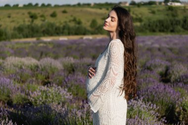 side view of pregnant woman with long hair standing with closed eyes in meadow with lavender