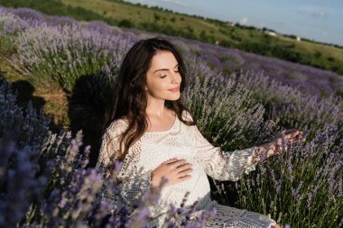 pleased pregnant woman in openwork dress sitting in lavender field