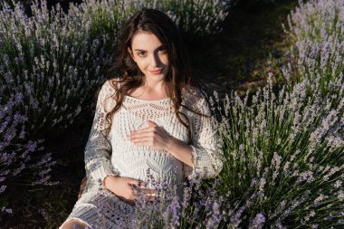 brunette pregnant woman looking at camera while sitting in meadow with flowering lavender
