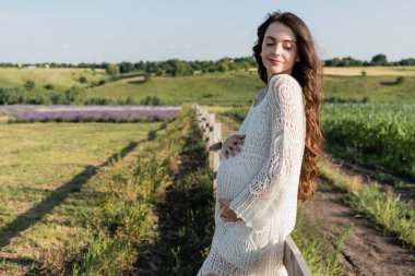smiling pregnant woman with hands on tummy standing in summer field
