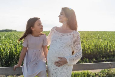 happy pregnant woman and girl in pink dress looking at each other near wooden fence in meadow