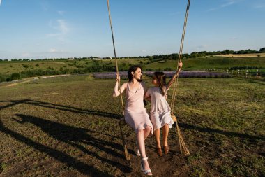 full length of woman and kid in summer dresses riding swing in meadow