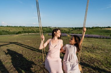 smiling mother and child riding swing in field and looking at each other