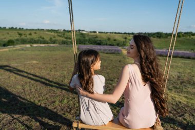 happy brunette woman looking at daughter while riding swing in meadow