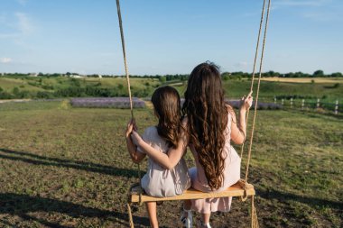 back view of long haired woman and girl riding swing in countryside clipart