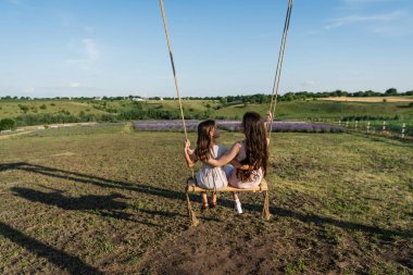 back view of brunette mom and daughter riding swing in meadow on summer day
