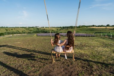 back view of brunette mom and daughter riding swing in meadow on summer day clipart