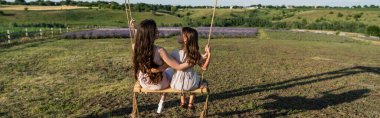back view of mother and daughter with long hair riding swing in field, banner