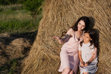 smiling woman pointing with hand while sitting on haystack with daughter