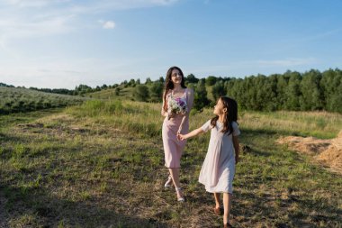 woman with bouquet holding hands with daughter while walking in field