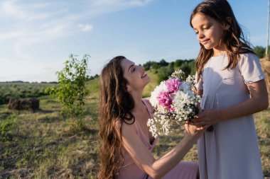 happy woman holding flowers near child in meadow on summer day