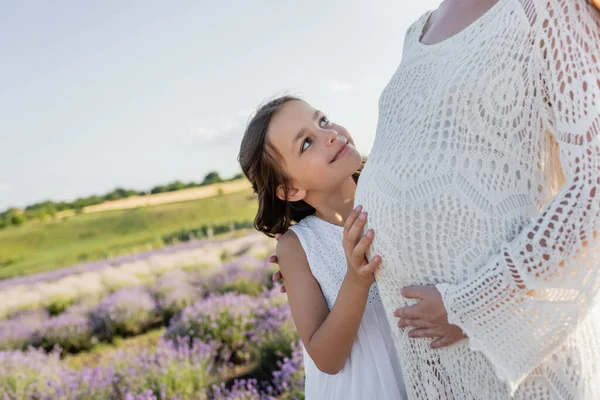 joyful child touching tummy of pregnant mother in blurred meadow