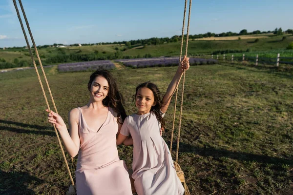 cheerful mom and daughter looking at camera while riding swing in meadow