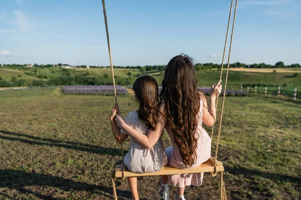 back view of long haired woman and girl riding swing in countryside