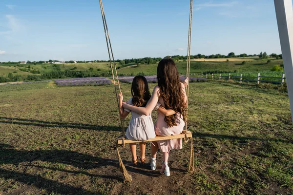 back view of brunette woman and child embracing and riding swing in field