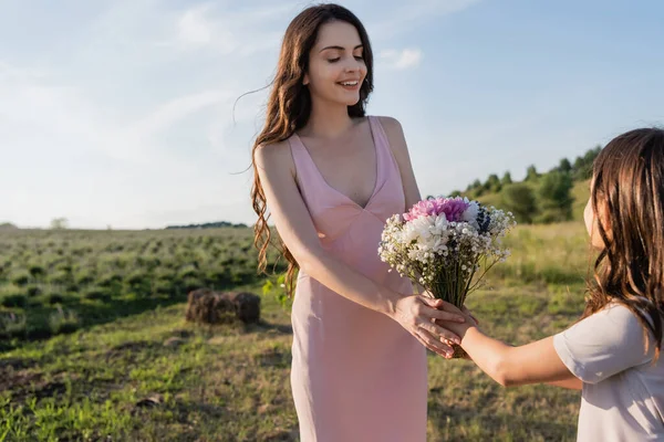 child presenting flowers to happy mother in pink dress in blurred field