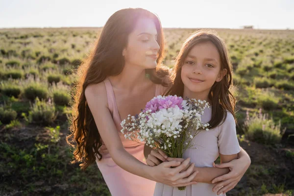 Brunette Woman Embracing Daughter Holding Flowers Field — Zdjęcie stockowe