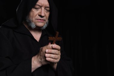 senior priest in dark hood looking at holy crucifix isolated on black clipart