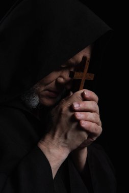 mysterious priest in hood obscuring face with crucifix while praying isolated on black clipart