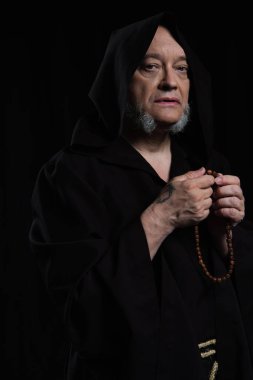 senior medieval monk with rosary looking at camera isolated on black clipart