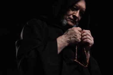 low angle view of monk in hooded robe praying with rosary isolated on black clipart