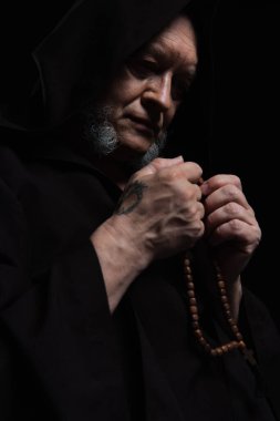 senior tattooed monk in hood holding rosary while praying isolated on black clipart