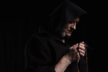 mysterious priest in hooded robe praying with rosary beads isolated on black clipart