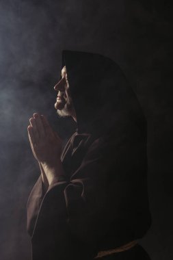 Kapüşonlu cüppeli keşişin dumanla siyah üzerine dua edişinin yan görüntüsü