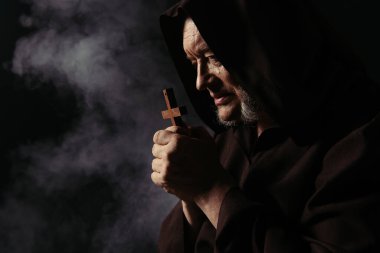 Siyah kapüşonlu cüppeli keşişin karanlık arka planda dumanla kutsal haçla dua edişinin yan görüntüsü