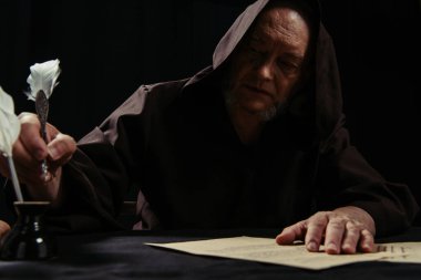 monk in hooded robe holding feather pen near inkpot and parchment with manuscript isolated on black clipart