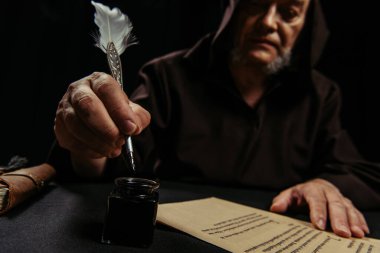 blurred monk holding feather pen near inkpot and ancient manuscript isolated on black clipart