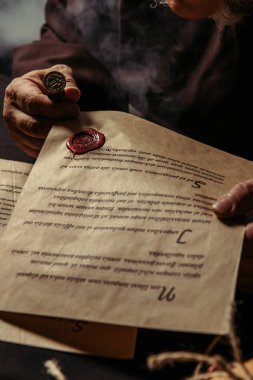 partial view of monk holding parchment with manuscript and wax seal on dark background clipart