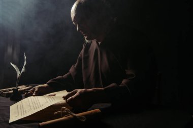 Siyah cüppeli ortaçağ keşişi karanlık arka planda dumanla antik el yazmalarını tutuyor.