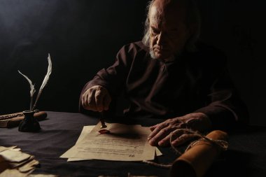 ancient monk stamping manuscript with wax seal near rolled parchment isolated on black clipart