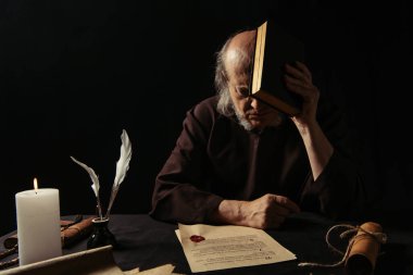 Yaşlı keşiş, siyah üzerine balmumu mühür yapıştırılmış İncil 'in yanında yüzünü gizliyor.